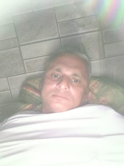A middle-aged man with short, graying hair and light skin is lying on a bed, covered by a white sheet. His head rests on a patterned pillow. Above him is a light-colored tiled wall. There is no verifiable information that the image was taken in Dhandhuka, India.