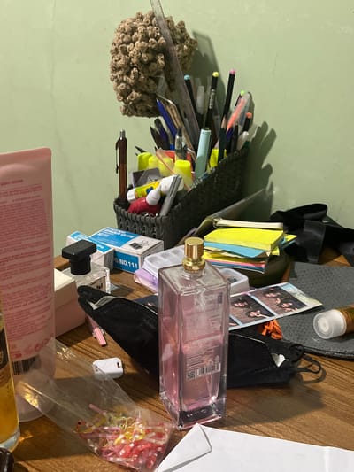 The image depicts a cluttered wooden desk in a room with a light green wall, likely located in Pasir Gadung, Indonesia. The desk is covered with various items including writing utensils in a dark basket, a pink lotion bottle, a perfume bottle, boxes of paper clips, sticky notes, a black face mask, and a small pile of colorful rubber bands in a plastic bag. A ruler is also visible amongst the pens. The overall impression is one of a disorganized workspace. There are no people or obvious actions occurring in the scene. The lighting suggests it may be indoors with artificial light.