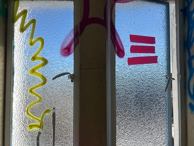 The image presents a close-up, interior view of a double-paneled frosted glass door or window, brightly illuminated by daylight from the exterior. The textured glass distorts the light blue of the sky visible through it, indicating clear weather.
Both glass panes are covered with distinct graffiti. On the left pane, a thick, vibrant yellow zig-zag line, resembling a stylized "ZZ," runs vertically down the glass. Below this, a faint, dark mark is also visible. The right pane features three horizontal, evenly spaced red rectangular shapes, which appear to be painted blocks or strips of tape.
A large, abstract spray-painted shape in pink or magenta is visible behind the central frame dividing the two panes, partially extending onto both glass surfaces. This shape is fluid and indistinct, suggesting part of a larger, unidentifiable design. White handles are present on both the left and right panes, indicating the functional nature of the window or door. Further graffiti, in the form of blue spray paint drips, is visible on a wall or frame just outside the right edge of the window.
The scene suggests an urban setting, likely within Dundee, United Kingdom, possibly an abandoned building or a public space, characterized by urban decay and graffiti art. The time of day is clearly daytime, given the bright, overhead lighting and clear sky. No readable text is present, only abstract painted shapes.