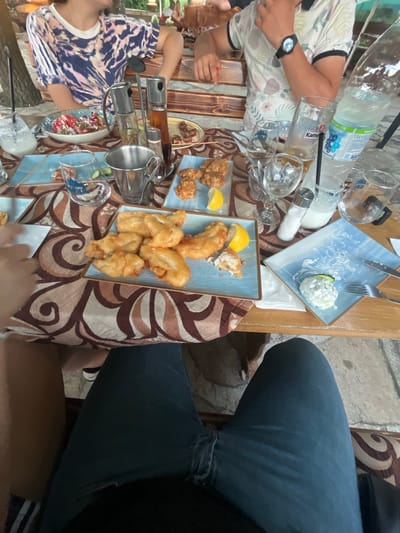 The image depicts a table setting with food and drinks, likely in an outdoor dining area given the presence of a stone floor and a glimpse of greenery in the background. The scene appears to be set during the daytime, with natural light illuminating the table.

Several subjects are visible. In the foreground, a person's arm and torso are shown, wearing a t-shirt with a colorful, abstract pattern. They are wearing a wristwatch on their left wrist. To the right, a person's legs are visible, clad in dark blue jeans.

On the table, there are several plates of food, including fried pieces of fish or chicken and a small salad. There are also glasses, some empty and some with remnants of drinks. A large plastic bottle with a label that appears to be for water is present, as is a salt shaker. A metal ice bucket is also on the table. The table itself is made of wood, and it's covered with a patterned tablecloth. Cutlery, including a fork and knife, are on a plate with some remaining food and a slice of cucumber.

The activity taking place is clearly a meal or a gathering around a table, with food and drinks being consumed. The overall impression is one of a casual meal, possibly at a restaurant or a picnic. There is a sense of relaxation and enjoyment.

Notable details include the water bottle, which has text that is partially visible but not fully legible. The setting is identified as Sushina, Bulgaria. The overall mood is relaxed and social.