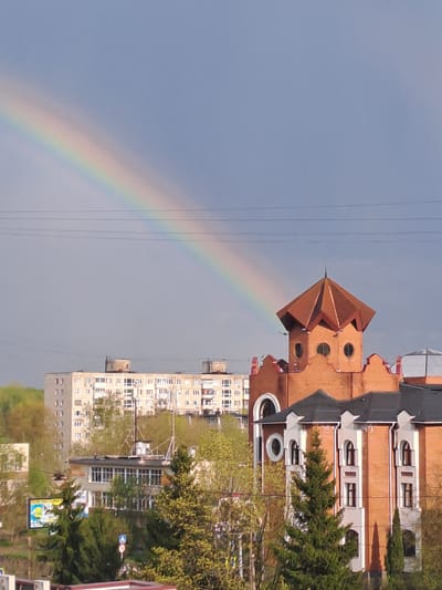 The image depicts a cityscape under a cloudy sky with a prominent rainbow on the left. A tall, ornate building with a copper-colored roof and circular windows is the main subject, and the rainbow appears to end near its spire. Other buildings, including apartment blocks, are visible in the background, along with trees and power lines. A billboard is seen on the lower right. It is likely the city of Yegoryevsk, Russia.