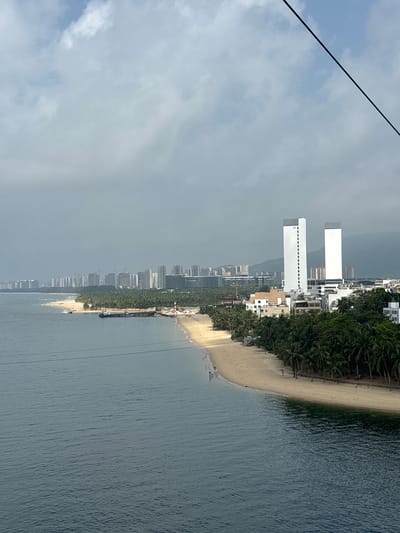This is a horizontal, medium-angle shot of a coastal city with a beach in the foreground. The water is a deep blue, and the sky is cloudy. The beach is lined with palm trees and buildings. In the distance, there is a cityscape with many tall buildings. There are a few people on the beach, and a few boats in the water. The image appears to have been taken from a high vantage point, possibly a cable car or a tall building. The location is the city of Xincun, China.