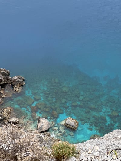 The image presents an elevated view of a tranquil coastal scene in Datça, Turkey. The majority of the frame, spanning the left and central areas, is dominated by the calm, blue waters of the sea.
The water on the far left appears a uniform, deep, and opaque blue, suggesting significant depth. Moving towards the right, the water becomes progressively clearer and lighter, transitioning into a vibrant turquoise. Through this translucent water, the rocky seabed is clearly visible, revealing numerous formations, boulders, and pebbles in shades of brown, grey, and green.
On the right side of the image, a rugged, light-colored rocky coastline descends into the sea. This shoreline is characterized by jagged rock formations and scattered stones. Several large, dark rocks are positioned along the water's edge, some partially submerged and appearing to have marine growth.
Sparse, hardy vegetation, including small green bushes and drier, gnarled shrubs, clings to the rocky terrain on the right. The scene is bathed in bright, natural daylight, indicative of a clear, sunny day. No human activity or visible text is present. The overall atmosphere is one of natural beauty and serenity.