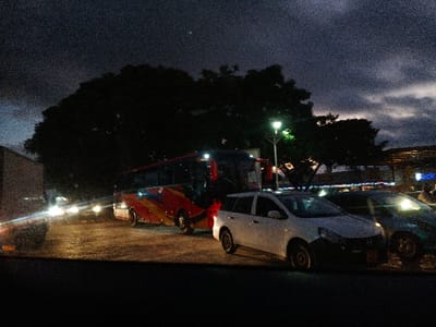 A night scene shows a bus with red and yellow designs, parked next to a white Nissan Livina. A green car is adjacent to the Nissan. A tall, dark tree stands behind the vehicles, with a street lamp shining through its branches. Building lights are visible in the background. A large truck is positioned on the left. The sky is dark, with clouds and minimal lighting.