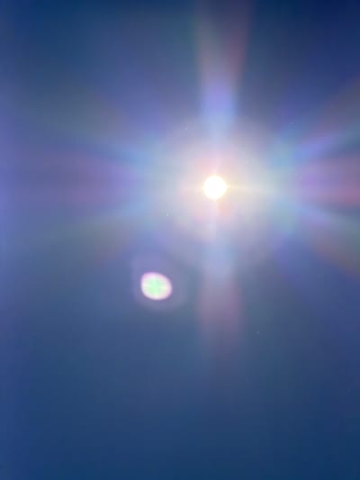 The image presents a bright light source, likely the sun, against a clear, deep blue sky. The sun, positioned slightly to the left of the frame's center, appears as an intensely bright white circular orb. Emanating from this central light are strong, multicolored light rays, creating a pronounced starburst or lens flare effect that stretches across a significant portion of the sky, displaying iridescent hues of purple, pink, and green.
To the right of the main light source, a distinct, out-of-focus, circular light anomaly is visible. This secondary light ghost is characterized by a blend of greenish and purple tones, indicative of further lens flare or internal reflections within the camera optics. Numerous tiny white specks are scattered across the entire blue sky, which could be attributed to dust on the camera lens or sensor, or perhaps very faint atmospheric particles.
The scene depicts a bright daytime setting with no clouds visible, suggesting clear weather. There are no human subjects, objects, or specific landmarks present to indicate a particular location within Bearpark, United Kingdom, aside from the provided context. The image captures the dynamic visual effects that occur when a camera is pointed directly at a powerful light source. No visible text is present in the image.