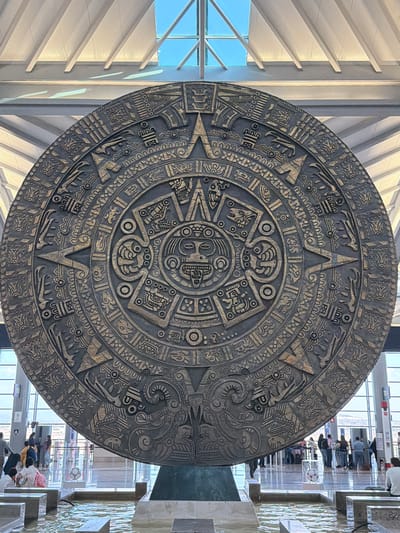 A large, circular, dark-toned replica of the Aztec Calendar Stone (Piedra del Sol), intricately carved with pre-Hispanic glyphs and a central facial figure, stands vertically on a dark pedestal within a shallow, rippling water feature. This scene is located in a spacious, modern indoor public facility in San Miguel Jaltocan, Mexico. The environment features a high, white vaulted ceiling with prominent structural beams and large skylights, allowing natural light to illuminate the space. In the background, beyond the water feature, large glass windows reveal a distant, arid landscape with low hills under a clear blue sky. Numerous individuals are visible in the mid-ground and background, standing and congregating on a light-colored, reflective floor, consistent with a busy public area such as an airport terminal. A white soccer ball is displayed within a clear case behind the water feature. Red vertical architectural elements are also present in the background. No specific interactions among individuals are discernible due to distance.