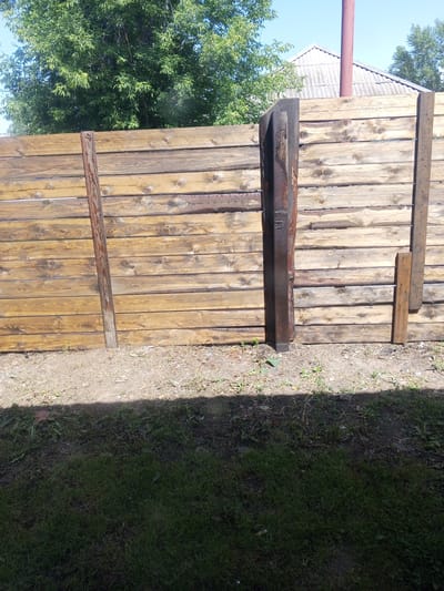 The image shows a weathered wooden fence with horizontal planks. A dark, vertical metal post is affixed to the fence on the left side, and a narrower wooden plank with nails is attached to the fence on the right side. The fence is situated in an outdoor environment with a patch of dirt and sparse vegetation in front of it, and lush green grass further in the foreground. Behind the fence, a large green tree with dense foliage is visible, and beyond that, a house with a corrugated roof and a red pipe. The sky is clear and blue. The scene is likely located in the city of Maslyanino, Russia, given the architectural style and vegetation.