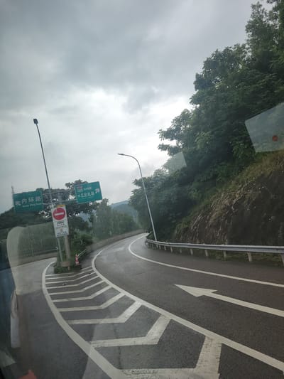 The image depicts a cloudy day in Yufengshan, China, with the perspective from inside a vehicle looking out at a curved road. The road has clear white lane markings, including directional arrows, and a metal guardrail runs along the side. To the left of the road, a steep, vegetated hillside rises, with lush green trees and foliage covering the rocky terrain.

Several signs are visible. One green sign with white text indicates "Inner Ring Road" (内环路) and shows a directional arrow pointing left. Another green sign, also with white text, indicates "G65 (渝邻高速)" and "Jiangbei Airport" (江北机场), with an arrow pointing towards the airport. Below these signs, a red and white circular sign with a horizontal bar signifies "No Entry" or a restriction, with additional text specifying time restrictions for "Passenger Vehicles" (客运车辆) from 02:00 to 05:00 and "Hazardous Chemicals" (危险化学品) from 00:00 to 06:00.

Streetlights are positioned along the roadside. The overall atmosphere is overcast, suggesting a potentially cool or damp day. The scene conveys a sense of travel and navigation, with the signs providing crucial information for drivers in the area. There are no people visible in the image, but the presence of road infrastructure and directional signs implies ongoing traffic and activity.