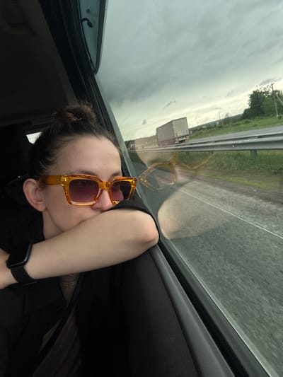 A person is resting their head on their arm while sitting in a vehicle, possibly a car, looking out the window. The person is wearing orange-framed sunglasses. A truck is visible on the road outside the window. The location is Kulaevo, Russia. The weather appears overcast. The person is wearing a black shirt and a smartwatch.