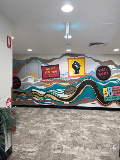 This media file presents an indoor hallway or corridor, brightly lit by several overhead circular lights and a vertical light fixture on the white left wall, which also features vents and access panels. The right side of the image is dominated by a colorful mural spanning the entire wall, while the floor is covered in a light-colored, patterned carpet.
The mural is a rich tapestry of abstract shapes and flowing lines, resembling a stylized landscape with interconnected rivers, streams, and rolling hills. The color palette includes shades of brown, teal, blue, and grey, accented with glittering gold. Embedded within this painted landscape are several framed messages and symbols. Towards the top right, a black rectangular sign with yellow text reads "BLACK WORK AHEAD" and "DO NOT OVERTAKE," accompanied by illustrative hands, a figure, and a traffic cone. Further left and slightly down, a yellow square features a black raised fist, symbolizing Black Power or solidarity. Below this, a red rectangular sign states "we are still here," with a camera icon.
Other elements within the mural include circular signs reading "BLACK LOVE" and "BLACK KNOWING," both with camera icons. Stylized shell-like objects are depicted along the winding streams, along with a dreamcatcher-like symbol. The overall themes of the muralโ"Black Love," "Black Knowing," "we are still here," and the raised fistโstrongly suggest a celebration and affirmation of Indigenous Australian culture and presence, pertinent to the Kelvin Grove, Australia location.
There is no activity taking place; the image captures a static interior. The artificial lighting indicates an indoor setting, with no discernable time of day or weather conditions. A red fire safety sign, partially legible as "ABE PONDER," is visible on the lower left wall.