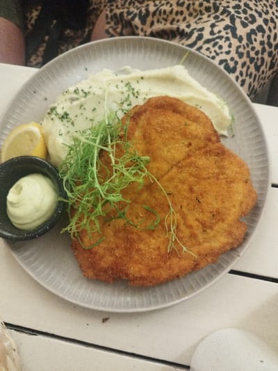 A plate of food on a white wooden table features a breaded cutlet, garnished with green sprouts. Next to the cutlet are mashed potatoes sprinkled with herbs, a lemon wedge, and a small black bowl filled with a creamy, pale green sauce. A person wearing a leopard print top is partially visible in the background. The plate is gray with a ridged pattern.