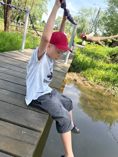 A child is hanging from a rope course over water. He wears a red baseball cap, a light gray T-shirt, and dark gray shorts, with black Crocs on his feet. He grips a purple rope ladder. The platform beneath him is made of wooden planks. The water reflects the green vegetation on the opposite bank. The location is Синьково, Russia.