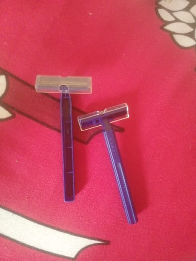 Two disposable blue razors with clear plastic protective covers are lying on a red textured surface. The razor on the left is oriented vertically, with its head pointing upwards. The razor on the right is angled to the upper right, with its head to the upper left. The red surface appears to be a cloth with a subtle pattern, possibly a rug or blanket, featuring cream-colored, stylized leaf-like shapes outlined in black. The image does not provide any information that could link it to Majemu, Nigeria or any specific city.