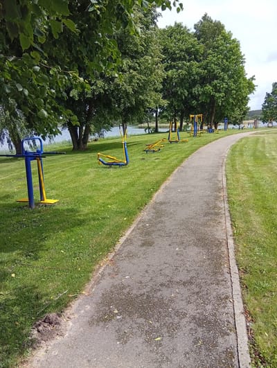 The image depicts an outdoor fitness area situated in Šaukėnai, Lithuania, adjacent to a body of water. A paved pathway curves through a grassy area dotted with various outdoor exercise machines, primarily in blue and yellow. In the foreground to the left, a blue and yellow leg press machine is visible. Further along the path, more exercise equipment is scattered across the lawn, including what appear to be bodyweight machines and pull-up bars. Large trees with green foliage line the background, providing shade. The body of water, likely a lake or river, is visible beyond the trees. The sky is overcast with clouds. The scene appears to be a public park or recreational space, with no people present. The ground on the left side of the path shows a disturbed patch of soil.