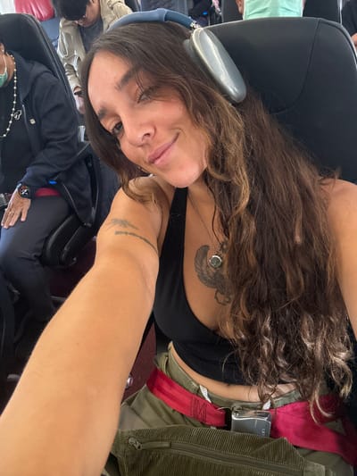 A woman with long brown hair and headphones sits on a plane. She wears a black tank top and has a tattoo on her chest. She also has a tattoo on her arm. A seatbelt is visible, secured with a metal buckle. Other passengers sit nearby, some wearing masks. The woman is looking at the camera with a slight smile.