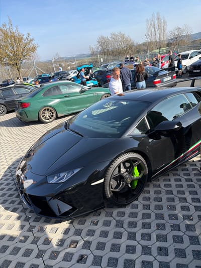 The image depicts an outdoor car gathering in Meuspath, Germany, under a clear, sunny sky. In the immediate foreground, a black Lamborghini Huracán is parked diagonally on gray interlocking concrete pavers. This vehicle features a green, white, and red stripe along its lower side panel and black multi-spoke wheels with neon green brake calipers. Behind the Lamborghini, a dark green BMW M2 coupe, equipped with bronze-colored multi-spoke wheels, is also parked.
The midground contains numerous individuals, primarily adults, standing and observing the parked vehicles. A man in a white collared shirt and glasses is visible, leaning over near the front of the dark green BMW. Further into the midground, a blue car has its rear hatch open, with a person in a blue shirt inspecting the interior. A black BMW E36 M3 with a large rear wing is also present, displaying a German license plate "LUF DF 238." Additional parked cars and groups of people are scattered throughout this area. The background features a sparse treeline, a wire fence, and distant rolling hills under a bright blue sky.