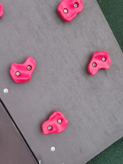 The image captures a close-up, high-angle view of a section of what appears to be a climbing wall or playground equipment, located in Nadym, Russia. The main surface is a dark grey, textured panel, likely made of rubber or a similar durable material.
Four distinct, irregularly shaped climbing holds, bright pink in color, are prominently featured on the grey panel. Each hold is secured to the panel by two visible silver-colored screws or bolts. The holds have a smooth, rounded surface, typical for children's climbing grips.
The grey panel itself seems to be composed of multiple sections, joined by visible metallic fasteners along its edges, particularly noticeable in the lower right portion of the frame. In the top left and upper right areas, a dark green, granular, and slightly uneven surface is visible, consistent with safety surfacing commonly used in playgrounds or outdoor recreational facilities.
The lighting is bright and diffuse, suggesting it is daytime, possibly under an overcast sky, as there are no harsh shadows. No individuals are present, and no specific activity is currently taking place, though the equipment is clearly designed for climbing and physical play. There is no visible text or branding anywhere in the image. The scene suggests a robust and well-maintained outdoor play area.