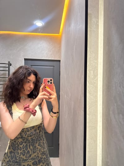 A young adult female is depicted taking a mirror selfie in a tiled room, likely a bathroom, located in Jrvej, Armenia. The subject has dark, wavy, medium-length hair with a center part and a slight pout. She is wearing a light yellow ribbed tank top beneath dark brown and tan leopard print denim overalls. Accessories include multiple necklaces (a light blue beaded choker, a pearl necklace, and a delicate chain with a pendant), a pink scrunchie and two thin gold bangles on her left wrist, and a black and gold bracelet with a thin gold bangle on her right wrist. Her fingernails are painted dark. She holds a smartphone with a red and white striped case and a small yellow clip in her right hand, angled towards the mirror.

The background features light gray/beige tiled walls with subtle texture. The white ceiling has a central circular recessed light fixture emitting white light and orange LED strip lighting along its perimeter. A dark metal towel rack is visible on the far left. A dark gray door with a silver handle is partially visible behind the subject. The mirror itself has a thin dark vertical border on its left edge.
