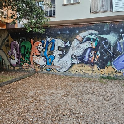 The image displays a section of a building facade adorned with vibrant graffiti art. The graffiti features a variety of stylized figures and symbols, including a purple, segmented creature with an eye, a green skull with crossbones, an orange character with an eye, and a large hand spraying a can of paint. The background of the graffiti is a dark, starry sky. To the left of the mural, a tree with dense green foliage partially obscures the upper part of the building. The building itself has multiple windows with white railings. In the foreground, a gravelly ground surface is covered with fallen leaves and a rough-hewn log lies horizontally. The overall scene suggests an urban outdoor environment, likely in the city of Girona, Spain, given the architectural style and the presence of graffiti.
