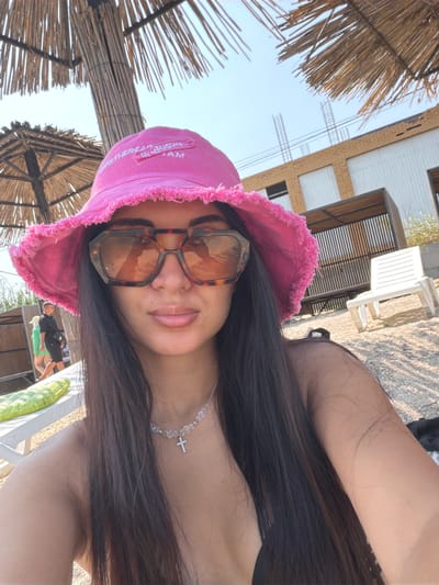 A medium shot captured from a low angle shows a woman wearing a pink bucket hat and large, tortoiseshell sunglasses. She is at the beach, as indicated by the sandy ground and a thatched umbrella overhead. Behind her, a partially constructed building with visible rebar stands against a clear sky. To the right, a white plastic lounge chair is positioned on the sand. In the background, two individuals are visible walking away from the camera, one in a black shirt and shorts, the other in a green top and shorts. A green inflatable object rests on a white lounge chair in the foreground. The woman is wearing a crystal necklace with a cross pendant.