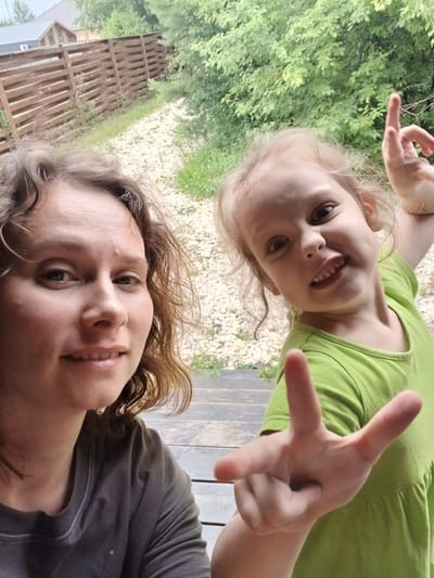 In Verkhneye Dubrovo, Russia, a woman and a young girl are captured in a selfie outdoors. The woman, with curly brown hair and a light complexion, is positioned on the left side of the frame, looking directly at the camera. She is wearing a dark gray t-shirt. To her right, a young girl with blonde hair is also looking at the camera, smiling and making a peace sign with her right hand. She is wearing a bright green t-shirt. The background features a wooden fence, lush green trees, and a gravel path, suggesting a rural or suburban setting. The overall atmosphere is casual and happy, indicative of a shared moment between the two individuals.