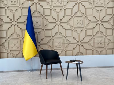 The image shows a Ukrainian flag mounted on a pole against a wall with a repeating geometric pattern in beige. To the right of the flag, a black leather chair with wooden legs sits next to a small wooden table with black legs. On the table are a pair of glasses and a sheet of paper. The location is likely an office or a building in Ankara, Türkiye, at coordinates 39.851, 32.827. The room has a white tiled floor and appears to be empty.  There is no one present in the image.