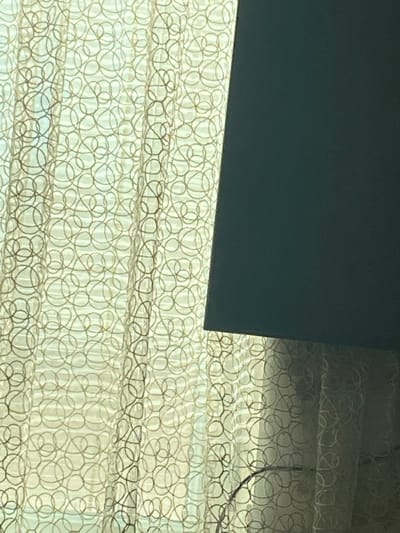 The scene depicts an interior setting within the city of Zamfir, Bulgaria. The primary subject is a sheer, light-colored curtain adorned with an embroidered or woven pattern of interlocking light brown concentric circles or ovals. Natural light emanates from behind the curtain, indicating a window or exterior opening. Obscuring the upper right and central right portions of the curtain is a dark, opaque, rectangular object, consistent in appearance with a television screen or monitor. Its lower edge extends diagonally downward from the upper right. In the lower right quadrant, a thin, light-colored, curved object, possibly a cable or part of a lamp stand, is visible in front of the curtain. No individuals or active interactions are present.