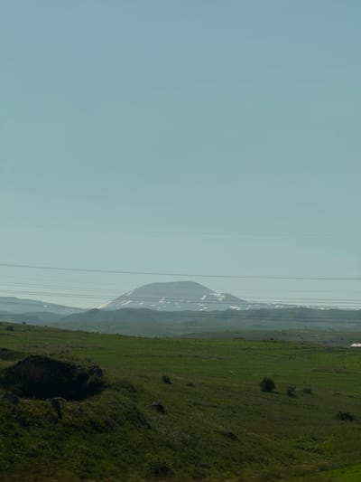 The image depicts a wide, open landscape under a clear, pale blue sky. In the foreground, a rolling green hill slopes downwards from left to right, covered in lush grass with scattered dark rocks and clumps of vegetation. A larger, dark, overgrown mound or structure is visible on the left side of the foreground. Further into the mid-ground, the landscape transitions to more distant, undulating green hills. A prominent mountain range, partially covered in snow, forms the backdrop. Several thin, horizontal power lines stretch across the mid-section of the image, obscuring parts of the mountain range. The overall impression is one of a natural, rural environment. The location is identified as being in Charentsavan, Armenia.