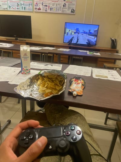 A person's hands are holding a black video game controller, with the person's legs, clad in camouflage pants, visible below the controller. The scene appears to be indoors, with desks and chairs, suggesting a classroom or training facility. On a desk in front of the person, there is an open bag of potato chips, a small pouch of sauce, and a carton of milk. In the background, a television screen displays a video game, and posters are mounted on the wall. The overall impression is of a casual break during a training session or lesson. It is not possible to determine the exact location in the city of Oredezh, Russia from the image.