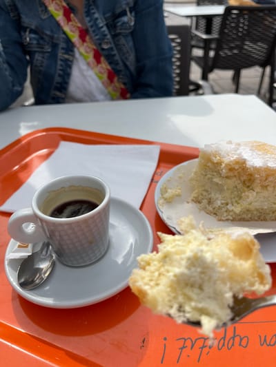 A close-up shot shows a coffee cup and a piece of cake, possibly brioche, on a tray, located in Palavas-les-Flots, France. The coffee cup, patterned with a waffle design, sits on a white saucer with a small spoon and a sugar packet. A portion of the cake sits on a separate plate, partially obscured. The tray is orange with the text "bon appétit!" visible. A person wearing a denim jacket with a patterned strap is visible in the background, sitting at the table. The background also includes a black metal chair. The scene suggests a café setting.