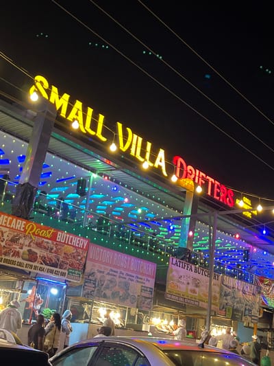 The image captures a lively nighttime scene in Kimorori, Kenya, showcasing a brightly lit commercial area with multiple food establishments. The main subject is a building adorned with numerous string lights in various colors, primarily blue and green, and prominent neon signage.
A large, diagonally oriented sign across the top reads "SMALL VILLA DRIFTERS" in illuminated yellow and red letters, with "GRILL" also visible. Below this, several food stall banners are displayed, advertising different butcheries and their offerings. Visible text includes "LECARE Roast BUTCHERY," "VICTORY ROAST BUTCHERY," "LATIFFA BUTCHERY," and "SAJANS BUTCHERY." The banners also list specific dishes like "Ugall Mukime Green Clips ha," "For The Dent Ceren. Fry Corken Chest & Fry," and "MBUZI CHONA, WET FRY DRY FRESHENTA." Contact numbers for orders, such as "0714 910 549" and "0719 181454 / 0746432609," are also present.
The scene is bustling with activity; several individuals, some dressed in white uniforms, are seen working at food stalls or interacting with customers. A car is partially visible on the right, suggesting roadside commerce. The dark sky indicates it is night, with the artificial lights creating a vibrant and energetic atmosphere. Overhead, utility lines are faintly visible against the dark backdrop.