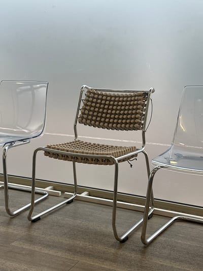 The scene depicts an interior view within Saint-Germain-en-Laye, France, showcasing three unoccupied chairs positioned in a row against a light-colored background, likely an opaque window or wall. The flooring is composed of dark brown, wood-patterned material.
From left to right:
1. A partial view of a transparent, clear plastic chair with a chrome-plated tubular metal frame, exhibiting a cantilever design. Only the left side of its backrest and seat is visible.
2. The central subject is a complete chrome-plated tubular metal chair, also featuring a cantilever design. Both its seat and backrest are fitted with tan or beige-colored textured pads. These pads are composed of numerous small, rounded, dome-shaped protrusions arranged in a uniform grid pattern, fastened to the chair frame with black cords visible on the sides.
3. A partial view of a third transparent, clear plastic chair, similar in design to the first, with its right side cut off by the frame.
A horizontal white bar or frame runs along the bottom of the light-colored background, approximately at the height of the chair legs. The environment appears clean and well-lit, with no individuals present. No actions or interactions are depicted.