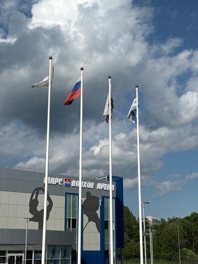 This image depicts the Volkhov Arena in Volkhov, Russia. Four flagpoles stand in front of the building, each flying a different flag. From left to right, the flags are white with a green emblem, the Russian tricolor (white, blue, and red), white with a red stripe, and white with blue and grey elements. The building's facade is gray with large black silhouette graphics of athletes, one appearing to be a hockey player and the other a figure skater. The text "ПАРС ВОЛХОВ АРЕНА" is visible on the building, indicating its name and location. The sky is partly cloudy with a mix of blue sky and grey clouds. Trees are visible in the background.