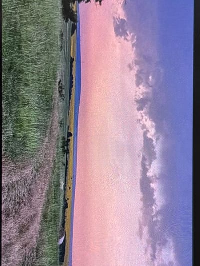 This media file captures a serene rural landscape, likely at sunrise or sunset, located near Wolverhampton, United Kingdom. The scene features expansive fields under a colorful twilight sky.

The sky transitions from a muted blue-purple at the top, down to a soft pink and orange gradient near the horizon. A prominent band of textured, darker clouds stretches horizontally across the mid-section of the sky.

Below the sky, the distant landscape reveals a thin strip of deep blue or purple land, possibly distant hills or another type of field. Beneath this, a wider field of vibrant yellow is visible, suggesting a crop like rapeseed in bloom.

The midground and foreground are dominated by a lush green field, likely grass or a young crop, with varying textures and heights. A dirt track or path with visible ruts winds through this green field, entering from the lower right and extending towards the center. To the far right in the midground, a large round hay bale rests in the field. On the left side, a cluster of dark trees or bushes partially frames the view.

The overall mood is peaceful and tranquil. There are no people visible, nor is any text present in the image. The photograph appears to be an image of a screen, as evidenced by a visible pixelation or screen-door effect throughout.