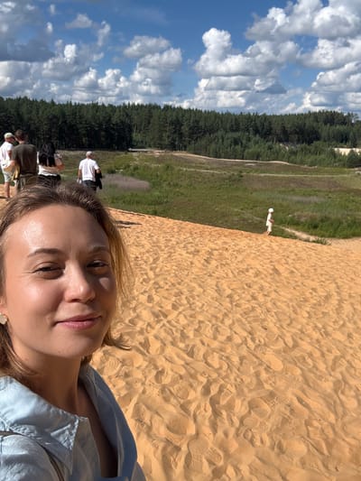 In the city of Luuska, Estonia, a selfie is taken in a sandy area with a forest in the background. A young woman with light brown hair, wearing a light blue collared shirt, smiles at the camera. The background features a sandy slope with footprints, a distant group of people, and a dense green forest under a bright blue sky with fluffy white clouds. A small child in white is visible in the middle ground, walking on the sand. The scene appears to be a sunny day in a natural, possibly recreational, outdoor location.