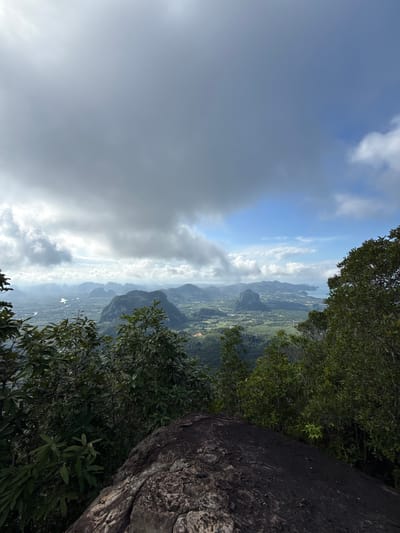 This is a landscape photograph taken from a high vantage point, overlooking a valley with karst formations and a coastline. The foreground is dominated by dark, textured rocks and lush green foliage from trees. In the middle ground, a sprawling valley unfolds, dotted with smaller hills, vegetation, and what appears to be a river or waterway. Further in the distance, more prominent, rugged karst mountains rise, some with visible structures or settlements at their bases. The horizon reveals a hazy coastline and the ocean. The sky above is a mix of dramatic, dark clouds and patches of clear blue sky with fluffy white clouds. The scene suggests a natural, tropical environment, likely captured during or after a period of rain due to the overcast conditions and potential haze. The location is identified as Ban Din Daeng Nui, Thailand, suggesting this viewpoint is from a mountain or elevated area within or overlooking this region. No people or animals are visible in the image.