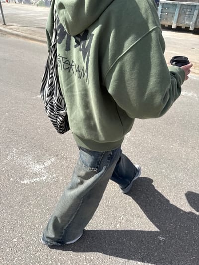 The image captures a person walking on an asphalt surface under bright daylight in Nes Ziona, Israel. The individual is viewed from behind and the right side, wearing an olive green hooded sweatshirt featuring black "STUSSY AMSTERDAM" text on the back, faded blue wide-leg denim jeans, and white athletic shoes. A black and white zebra-striped tote bag is slung over the left shoulder. The person holds a black disposable coffee cup with a lid in their right hand. Distinct shadows are cast on the light gray pavement, which exhibits faint white line markings. In the background, a light blue industrial skip is visible on the right.