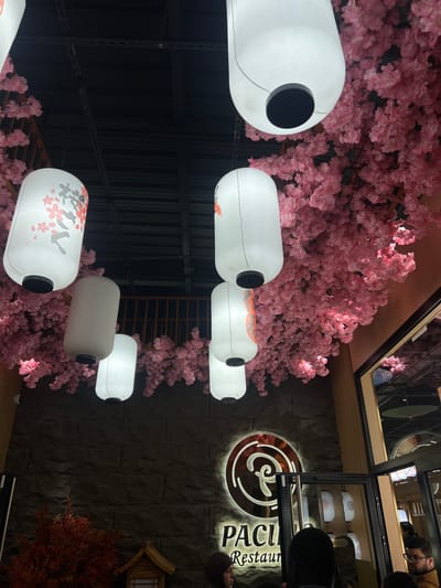 The image presents the interior of "PACIFIC Restaurant" in Clamart, France, characterized by a detailed decorative theme. An artificial canopy of dense pink cherry blossom foliage covers large sections of the dark, exposed industrial-style ceiling, from which multiple white cylindrical lanterns are suspended. Several lanterns feature black bases and tops; at least three display red and grey/black Japanese characters or floral designs. One lantern in the upper center exhibits a distinct black circular opening at its base.
A dark grey, textured stone-block wall serves as a backdrop on the left and center. Mounted on this wall is an illuminated sign for "PACIFIC Restaurant," with a stylized 'P' logo enclosed within a circular emblem, emitting a warm orange light. In front of the stone wall, in the lower left, a dense bush with reddish-brown foliage is positioned beside a small, traditional Japanese-style wooden decorative structure with a tiered roof.
On the right side, vertical light-colored wooden slats frame an entrance or large window, reflecting parts of the overhead cherry blossom decor and an ornamental Japanese umbrella-like fixture. Several individuals are present in the lower right foreground, positioned near this entrance. Their faces are largely obscured; one man on the far right is visible with a beard, glasses, and a light brown jacket, looking towards the right. The back of a person's head with dark hair occupies the immediate foreground, partially obscuring the restaurant sign. A person wearing a dark head covering is partially visible on the far left. No specific actions or interactions among the individuals are discernible beyond their presence in the establishment.