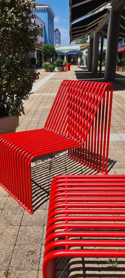 This image depicts a modern, bright red, tubular metal bench placed outdoors on a paved area. The bench has a distinctive zig-zag design, with multiple parallel tubes forming the seating surface and backrest. The scene appears to be a pedestrian area, possibly a shopping street or plaza, given the presence of shopfronts and awnings visible in the background. The architecture of the buildings suggests a contemporary design, with some featuring colorful facades.
In the background, several people can be seen walking, interacting, or browsing. One shop prominently displays the "Tommy Hilfiger" logo, providing a specific location cue. Lush green trees and potted plants are also visible, adding to the outdoor ambiance. The sunlight casts strong, striped shadows from the bench onto the pavement, indicating it is daytime, likely midday or early afternoon due to the sharp shadows. The sky is clear and blue, suggesting fair weather.
The overall impression is one of a pleasant, sunny day in a well-maintained urban or commercial outdoor space, likely in Aiello del Friuli / Dael, Italy, as indicated by the location context. The vibrant red of the bench contrasts with the neutral tones of the paving and the natural greens of the foliage, making it a focal point of the composition.