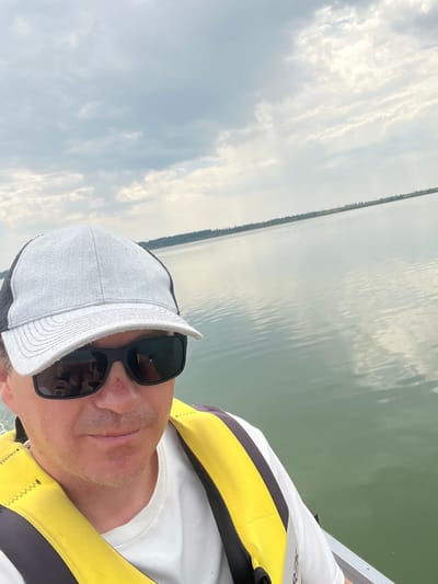 A selfie taken from a low angle shows a man wearing a grey baseball cap with a black mesh back, black sunglasses, and a yellow and black life vest over a white t-shirt. He is on a body of water, possibly a lake, with a shoreline visible in the distance. The sky is overcast with clouds. The water is calm with reflections of the sky. The location is identified as Beregovoye, Kazakhstan.