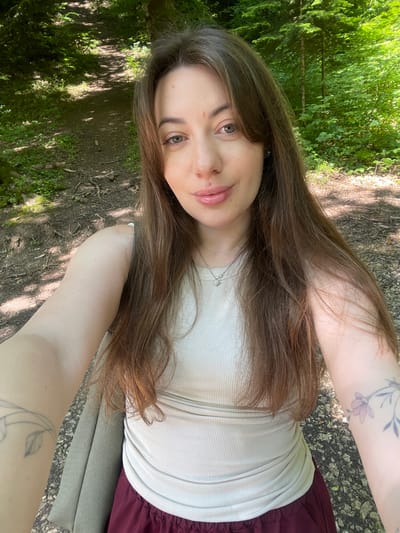 In Guzeripl', Russia, a young woman with long brown hair, wearing a beige ribbed tank top and a maroon skirt, takes a selfie outdoors. She has a delicate necklace with a star pendant. Her right arm is visible, showing a tattoo of a flower. She is positioned in a forest clearing with a dirt path winding through it, shaded by lush green trees. Sunlight filters through the leaves, dappling the ground. The overall scene suggests a peaceful nature walk.