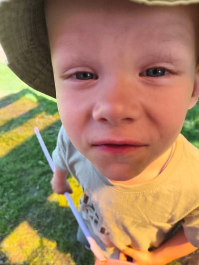 A young child with fair skin, blue eyes, and a serious expression is looking directly at the camera. The child is wearing a light-colored bucket hat and a light-colored t-shirt. They are holding two light purple objects, likely glow sticks. The background shows green grass, suggesting an outdoor environment, potentially a yard or park. The location is given as Chernaya, Russia, though there is no direct visual information to confirm the city. The lighting suggests it is daytime. The perspective is from a low angle, as if the photographer is kneeling or sitting.