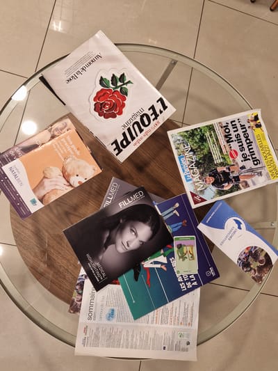 The image shows several magazines and brochures spread out on a round glass-top table with a wooden frame. The table is situated in a space with light-colored, tiled flooring, indicative of an indoor environment, possibly a store or a waiting area. The lighting suggests a well-lit indoor space. One magazine prominently features a large red rose on its cover with the title "Au nom de la Rose." Another magazine has the word "MAGAZINE" in large, stylized letters, with a picture of a man cycling. A third magazine has the title "Le Parisien" and is about the Tour de France. There are also other publications visible, including a brochure with a teddy bear and text related to pregnancy, and a magazine titled "FILLMED LABORATORIES." The overall impression is a collection of reading materials laid out for display or perusal. The question about the location in Antony, France, cannot be answered from the visual information provided in the image.