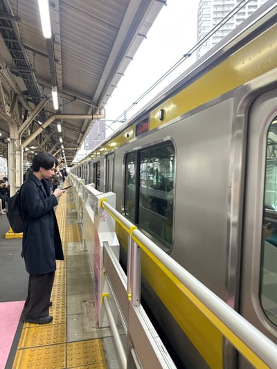 A train at a station, angled upward, shows the roof and side of a silver train with a yellow stripe. A person in a dark coat stands on the yellow-textured platform, looking at their phone. The platform is covered by a metal roof with fluorescent lights. Tall buildings are visible beyond the platform roof. Other people wait further down the platform.