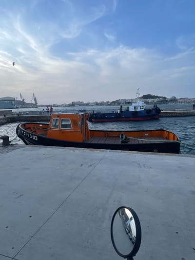 An orange and black tugboat, bearing the identification number 5 CT-43-03, is moored to a concrete quay. The boat has a wooden deck and a cabin structure with windows. Behind it, a larger blue and white tugboat is also in the water. In the background, the city of Cartagena, Spain, is visible with its buildings and a hill. Cranes suggest an industrial or port area. The sky is partly cloudy with a bird flying in the upper left quadrant. A circular mirror, likely from a vehicle, reflects buildings and a street scene in the lower right corner of the image. The overall scene depicts a waterfront area in a Spanish port.
