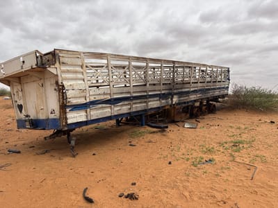A wide shot of a damaged, white and blue livestock trailer abandoned in the sandy terrain of Um Laba, Sudan. The trailer is heavily weathered and rusted, with visible signs of disrepair. The wooden slatted sides of the trailer are open, suggesting it may have been used for transporting animals. The ground around the trailer is covered in sand, with scattered debris and sparse vegetation. The sky is overcast, with heavy clouds. There are no people visible in the image.