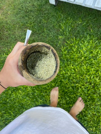 A top-down, eye-level photograph captures a hand holding a traditional South American gourd (mate) filled with yerba mate and a metal straw (bombilla). The hand is positioned over a bright green lawn, and a person's bare feet are visible in the background. A section of a white structure is visible in the upper left corner. The person wears a white shirt. The location is likely in Gerli, Argentina, based on the cultural association of yerba mate with the region.