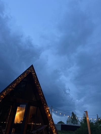 The image depicts a low-angle shot of a building structure with string lights illuminating its edges against a backdrop of a cloudy, blue evening sky. In the upper right, a wooden pole supports additional string lights stretched over a darker building partially hidden behind trees. The photograph was taken at night. The location given is Малеевка, Russia. No people are visible in the image.