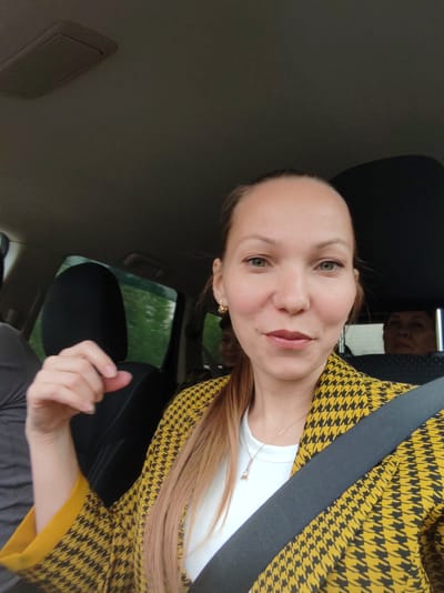 A woman with fair skin, green eyes, and light brown hair pulled back in a ponytail is seated in the driver's seat of a car, wearing a white t-shirt, a yellow and black houndstooth blazer, and a seatbelt. She is looking at the camera, and her right hand is raised. In the backseat, two other people are visible; their faces are partially obscured by the front seats. The car appears to be in motion. The exterior seen through the windows suggests a wooded area or park. Based on the query, the location is Pazhga, Russia.