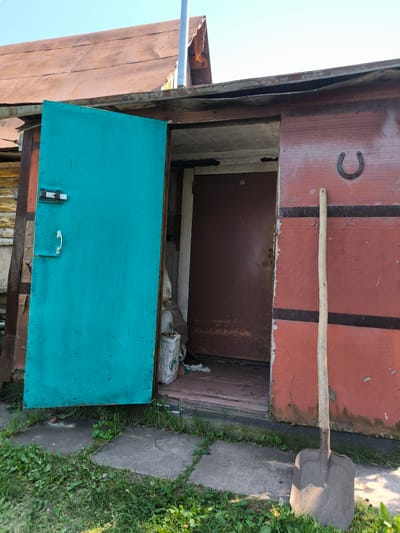 The image depicts a rustic wooden structure with a red-painted exterior and a slightly rusted, copper-colored corrugated metal roof. A large, bright turquoise metal door stands ajar on the left side of the structure, revealing a glimpse of the interior. To the right of the entrance, a dark brown door is visible. Mounted on the red wall above the dark brown door is a black horseshoe. Leaning against the red wall, to the right of the entrance, is a shovel with a wooden handle and a metal blade. The ground in front of the structure is covered in patches of grass and concrete pavers. The sky above is clear and blue. The scene appears to be in an outdoor setting, potentially a shed or outbuilding, in Maiskii, Russia.